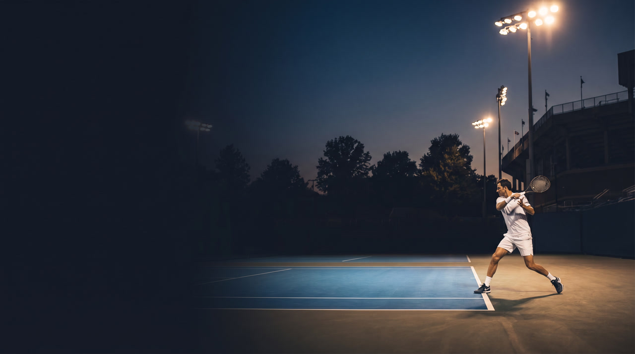 Lindner Family Tennis Center Center Court at the Cincinnati Open during an evening session, blue Laykold hard court warm under stadium floodlights, Midwestern tree line and stadium architecture in shadow under a deep navy Ohio summer sky