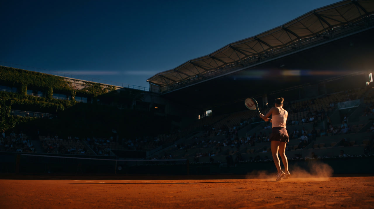 Court Philippe-Chatrier at Roland Garros in late Parisian afternoon, rust-orange terre battue clay glowing under western light, ivy-clad upper stands shadowed against a navy evening sky